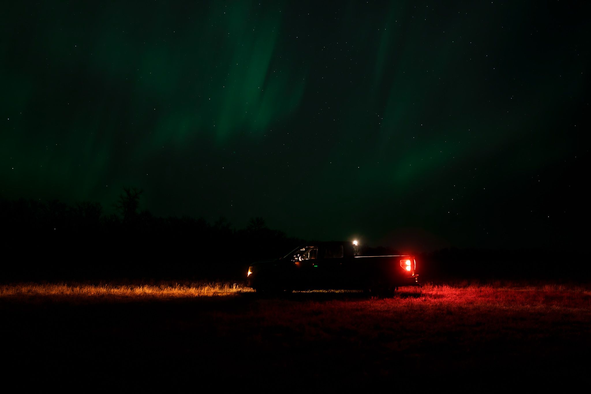 Green aurora borealis dances across a dark night sky over a snowy winter landscape with red lights in the distance.