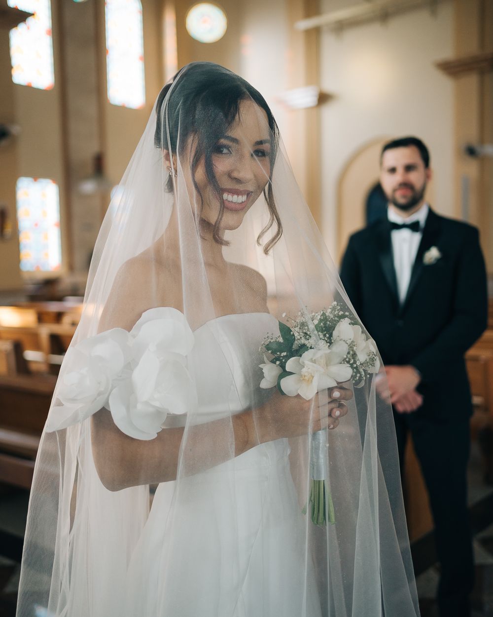 noiva sorrindo com véu e buquê durante cerimonia de casamento na igreja com noivo ao fundo