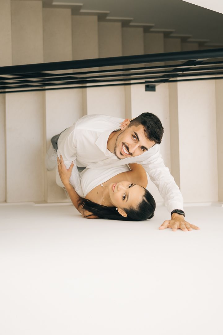 casal de noivos em pose artística e descontraída no chão, com noivo inclinado sobre a noiva sob uma estrutura de escadas modernas