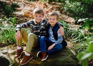 Two pairs of brown leather boots sit on a rustic log surrounded by autumn leaves in a natural outdoor setting.
