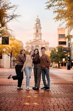 Four people in casual fall attire pose playfully on cobblestone street with historic church building in background at sunset.
