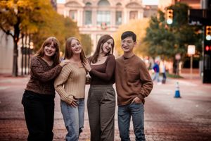 A group of four friends stand together smiling on a brick street with autumn trees and historic buildings in the background.