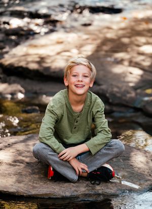 Child in olive green henley shirt sitting on rocks near a stream in dappled sunlight.