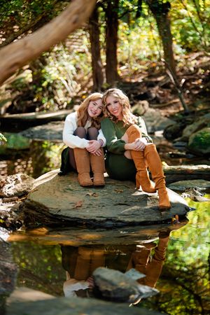 Two people relax on a large rock by a reflective pond in a serene forest setting.