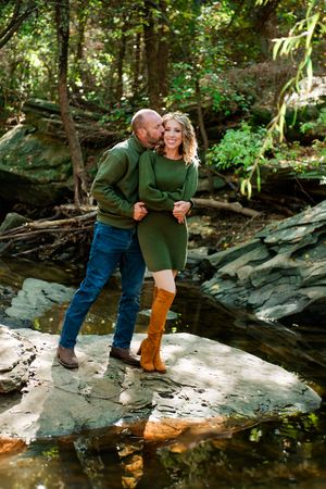 A couple in matching green sweaters share an intimate moment while standing on rocks by a forest stream.