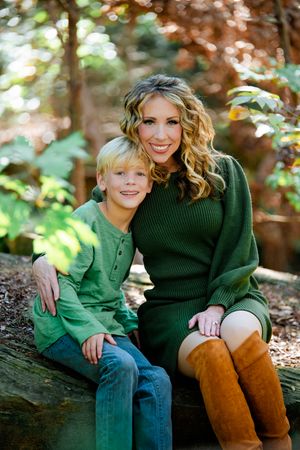A parent and child sit together on a fallen log in autumn woods wearing green and brown sweaters and boots.