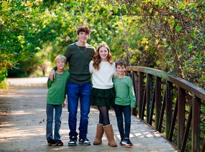 A family wearing coordinated green outfits poses together on a wooden bridge in a sunlit outdoor setting.