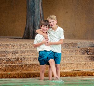 Two siblings sit together on stone steps near a pool wearing matching outfits on a sunny day.