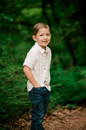 A young child in a white shirt and jeans stands playfully in a wooded outdoor setting with lush green foliage.