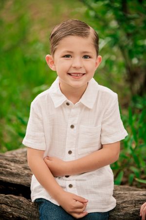 A child in a white button-up shirt poses outdoors on a fallen log with a natural green background.