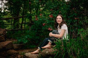 A person sits on a stone wall surrounded by lush greenery and red flowers in a garden setting.