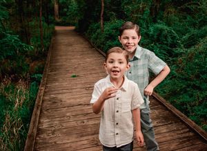 Two children make silly faces while standing on a wooden boardwalk trail surrounded by lush green forest.