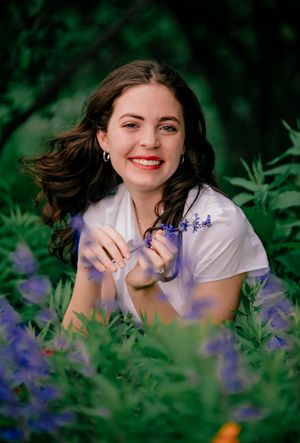 A person lying in a field of purple wildflowers smiles while surrounded by lush green foliage in a summer portrait.