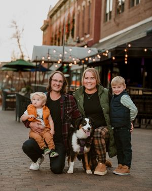 A family group sits on brick steps with their small dog while string lights glow in the background at dusk.