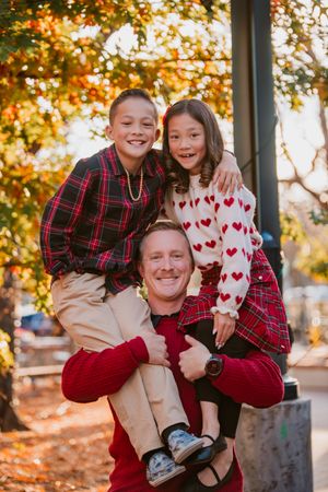 Candid family moment captured outdoors with fall foliage backdrop wearing matching red and plaid outfits.