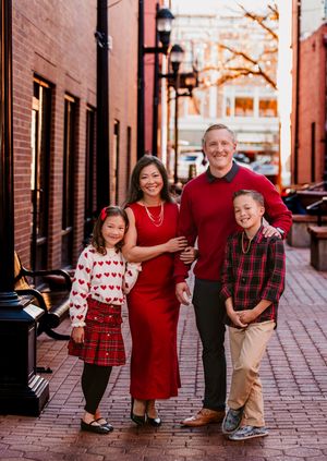Family wearing red and plaid outfits poses together on brick sidewalk for holiday photos.