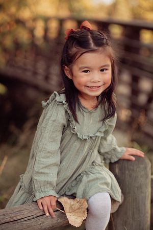 Child in mint green dress sits on wooden fence holding autumn leaf while enjoying a sunny fall day outdoors.