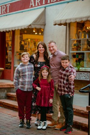 A family poses together in front of a brick storefront decorated for the holidays with children wearing festive attire.