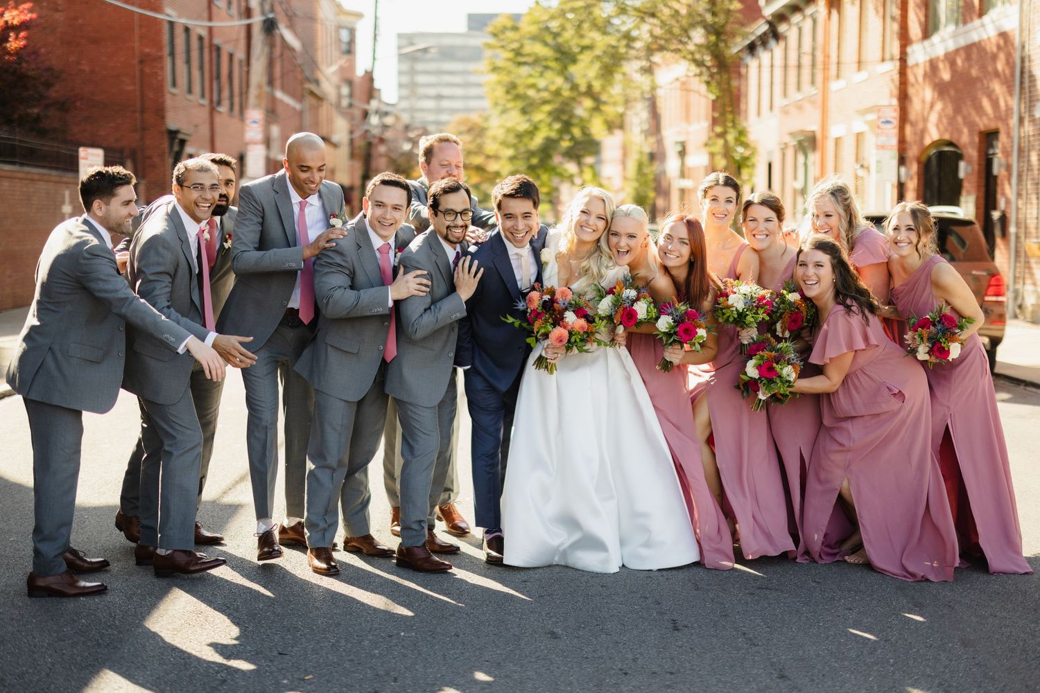 Wedding party in gray suits and pink dresses posing on urban street.