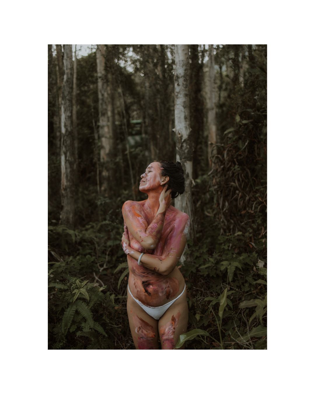 Artistic nude figure poses expressively in a moody forest setting with dramatic lighting and body paint.