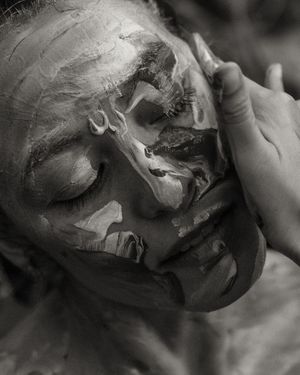 Black and white artistic portrait showing a face covered in thick, textured paint or clay with closed eyes.