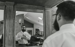 A black and white photograph shows a bearded person adjusting their shirt collar while looking in a bathroom mirror.
