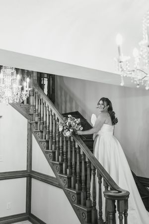 A bride in a white wedding dress poses elegantly on a classic wooden staircase with ornate railings.