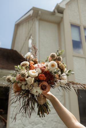 Beautiful autumn-themed wedding bouquet with orange and white flowers held up against a light blue sky.