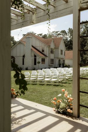 White wedding venue with rows of folding chairs set up on a lawn, viewed through a pergola with climbing vines.