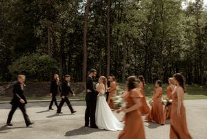 Wedding party walking through a wooded outdoor path with bridesmaids in rust-colored dresses.
