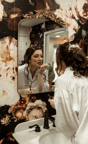 Wedding preparation scene in bathroom with ornate floral wallpaper and vintage-style mirror.