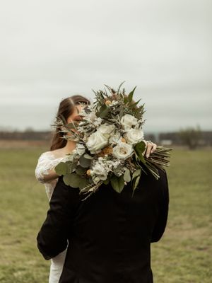 A beautiful white and green bridal bouquet held against a cloudy sky and open field.