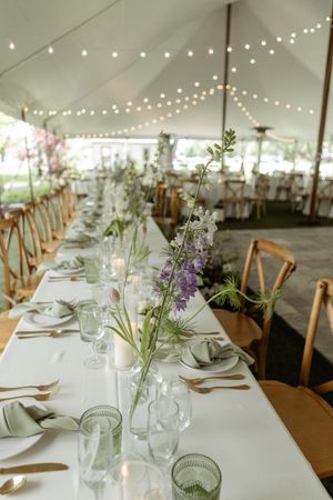 Elegant wedding reception tables set with white linens, natural centerpieces, and string lights in a spacious tent pavilion.