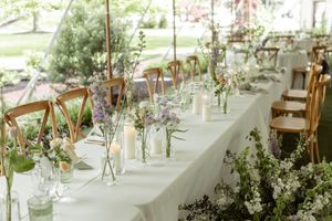 A wedding reception table decorated with delicate wildflowers in glass vases and wooden chairs in an airy venue.