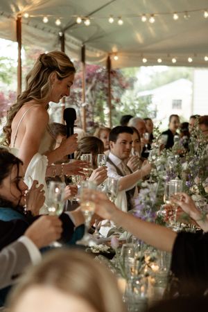 A wedding reception under a tent with string lights as guests raise glasses for a toast during a speech.