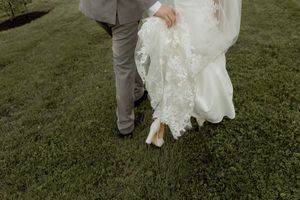 A white wedding dress with ruffles and a gray suit, holding hands and walking across a green grass lawn.