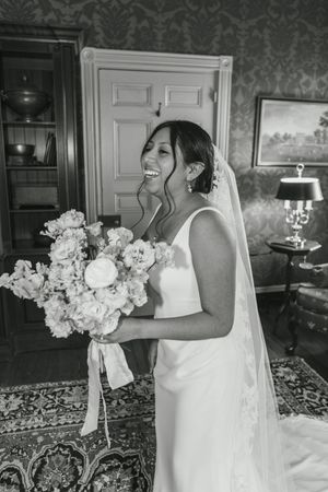 A bride in a white wedding dress holds a beautiful bouquet of white roses and flowers in a decorated indoor setting.