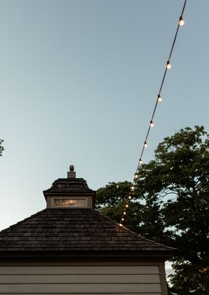 String lights hang between wooden posts against a clear blue sky with a black shingled roof visible.