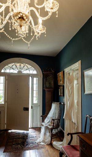 Elegant entryway with crystal chandelier, dark teal walls, hardwood floors and white wedding dress displayed near a rocking chair.