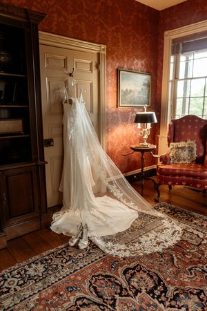 An elegant white wedding dress and veil hanging on a door in a luxurious room with red damask wallpaper and oriental rug.