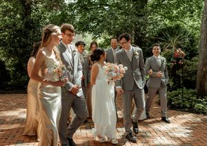 Wedding party walks and laughs together on a brick pathway surrounded by lush greenery on a sunny day.