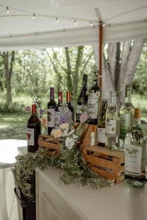 Rustic wooden crates display wine bottles with floral arrangements under a white tent canopy at an outdoor event.