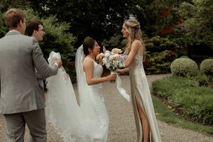 Two people in wedding dresses with white bouquets share a moment on a garden path while others look on.