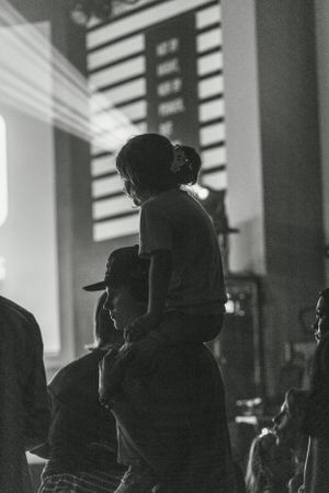 A silhouetted crowd watches as a child sits on shoulders during an event with a scoreboard visible in background.