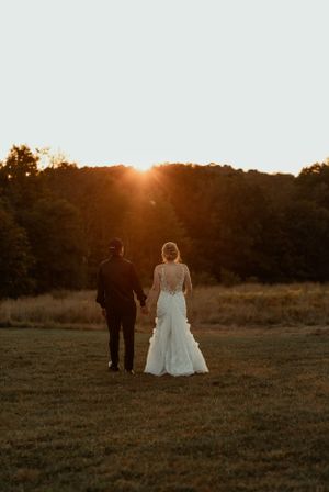 Newlyweds walking through a golden field at sunset with trees in the background.