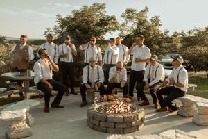 A group of groomsmen in suspenders gather around a stone fire pit during golden hour.