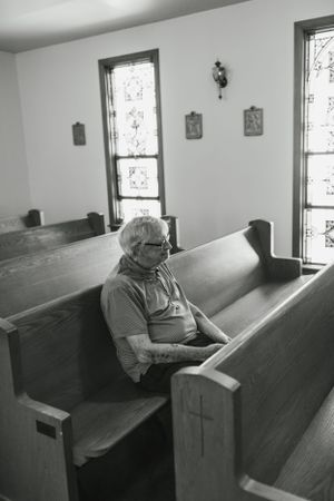 A black and white photo of someone sitting alone in an old wooden church pew.
