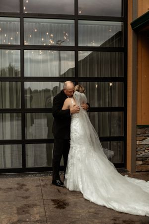 Wedding couple embraces in front of modern glass window wall with string lights.