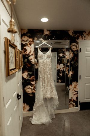 A delicate lace wedding dress hangs elegantly against a dark floral wallpaper in a bridal suite.