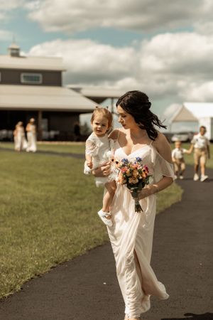 A bride in flowing white dress walks along a path carrying a coral bouquet while holding a small child.